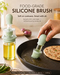 A silicone oil brush with a pale mint green silicone head and clear body is shown being used to grease a black frying pan with oil, alongside a matching oil dispenser bottle. The scene is set in a warm kitchen environment with a flatbread on the pan, green chilies, and onions in the background, with text overlays reading 'FOOD-GRADE SILICONE BRUSH'.